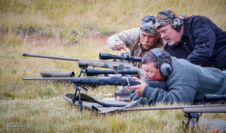 Three men aiming rifles at an outdoor shooting range. One man is prone, two others are standing and observing.