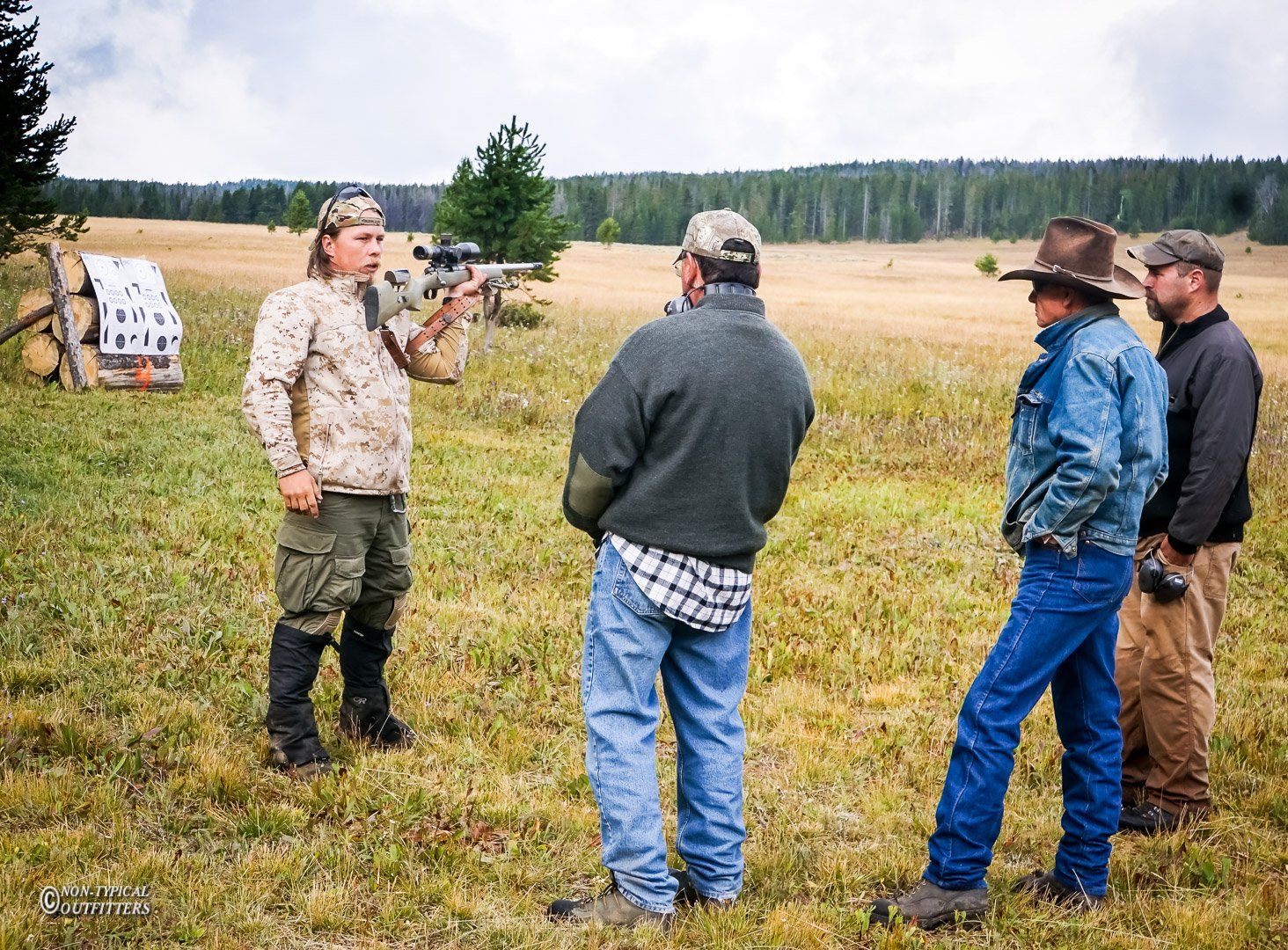 A man in camo points a rifle, speaking to a group of men in a grassy field.