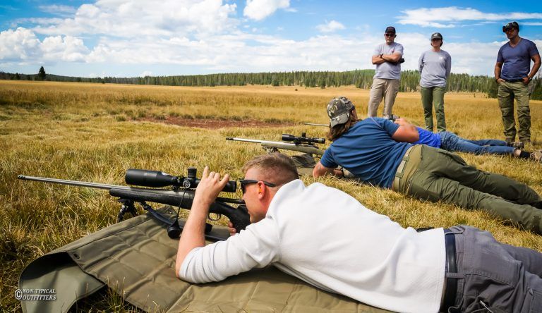 People shooting rifles at a range on a sunny day. Others watch from behind.