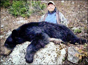Man poses behind a large, dead black bear lying on a rock outdoors.