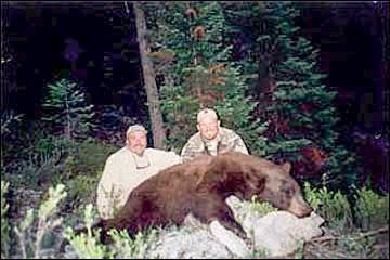 Two men pose with a dead black bear in a forest setting.