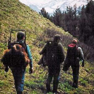 Three people hiking up a hillside. One carries a large pack, others have backpacks. Mountains in background.