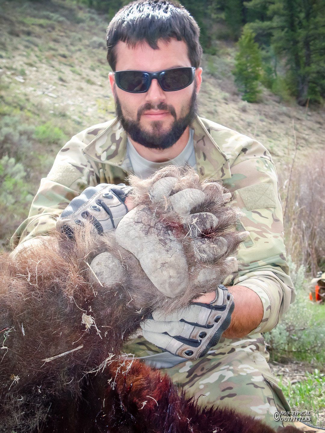 Man in camo holding a large bear paw outdoors; paw covered in dirt.