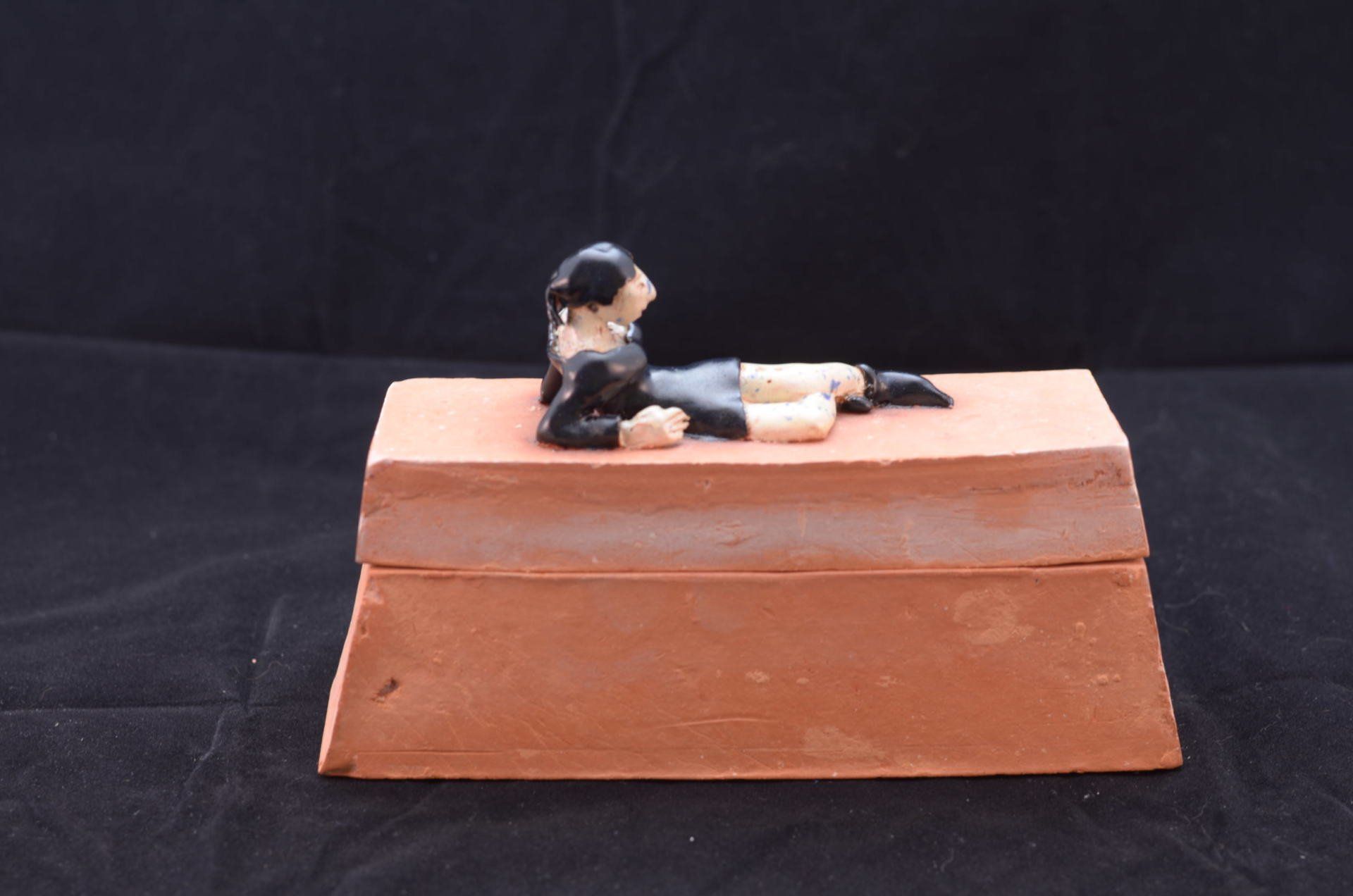 Een klein figuurtje met zwart haar en een zwarte outfit ligt op zijn zij bovenop een rechthoekig, terracottakleurig kleiblok.