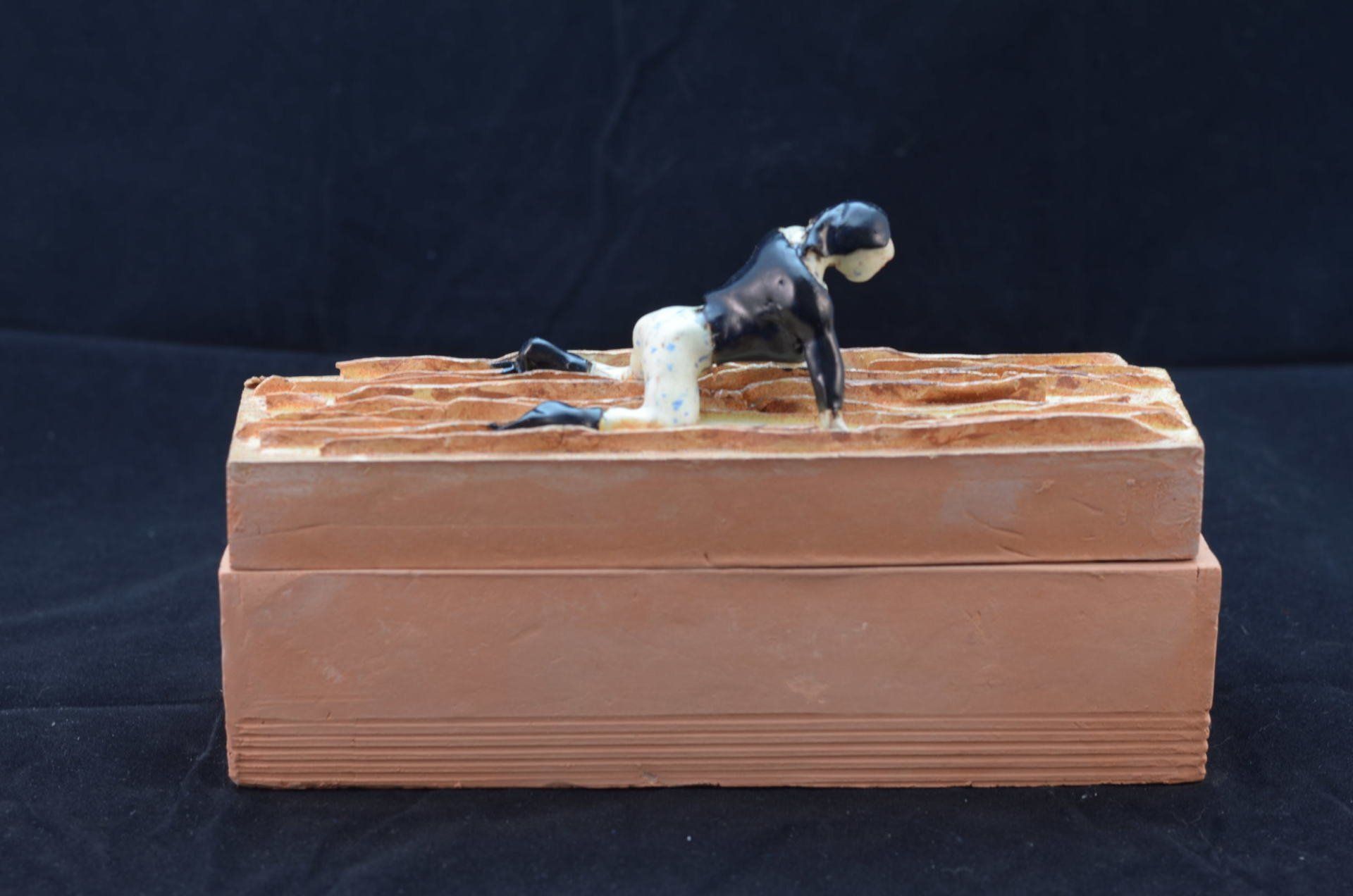 Een knielend figuurtje met donker haar en een witte broek staat bovenop een rechthoekig, gestructureerd terracotta blok.