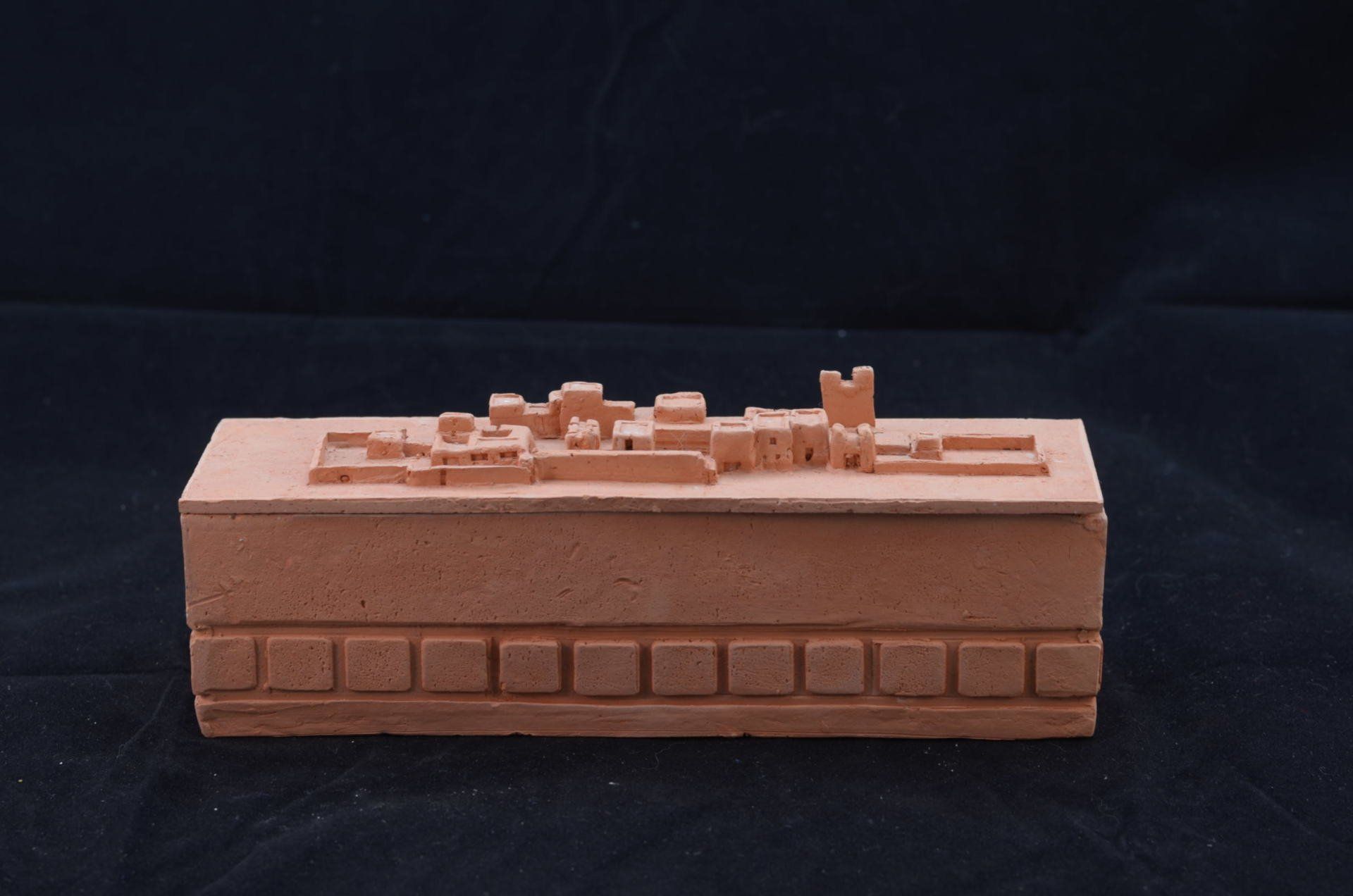 Een object in terracotta-kleur met een miniatuur uitgesneden kashba bovenop en een rij vierkante blokken aan de basis.