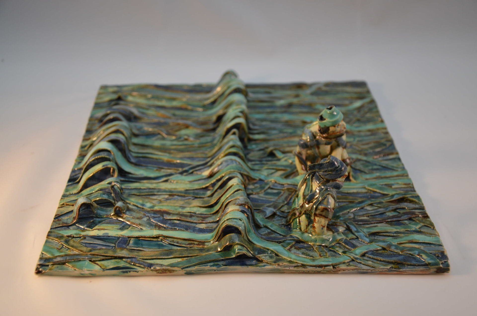 Een keramische tegel in tinten turquoise en blauw is voorzien van een figuur die in een gebeeldhouwde, golvende zee staat.