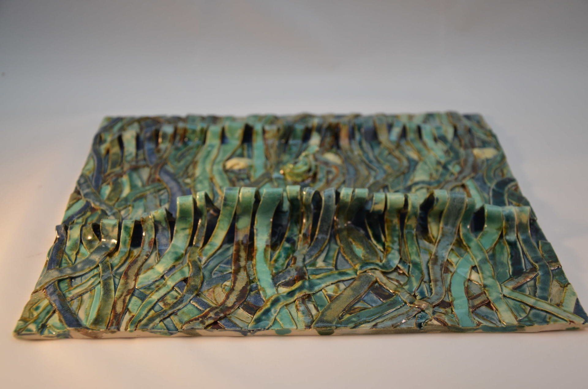 Een vierkant keramisch kunstwerk met golvende, gestructureerde, turquoise en groene strepen die op zeewier lijken.