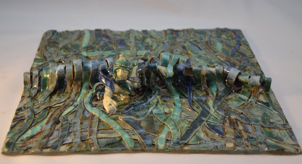 Een reliëfsculptuur van keramisch materiaal in groen en blauw, met een textuur, toont een centrale, ruggengraatachtige structuur omgeven door lintvormen.