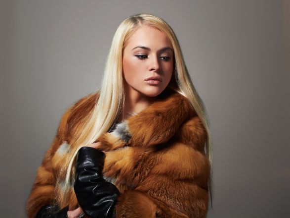 Modella bionda con cappotto di pelliccia marrone e guanti neri, in posa davanti a uno sfondo grigio in studio.