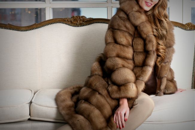Una donna con un cappotto di pelliccia marrone è seduta su un divano color crema in un luminoso soggiorno.