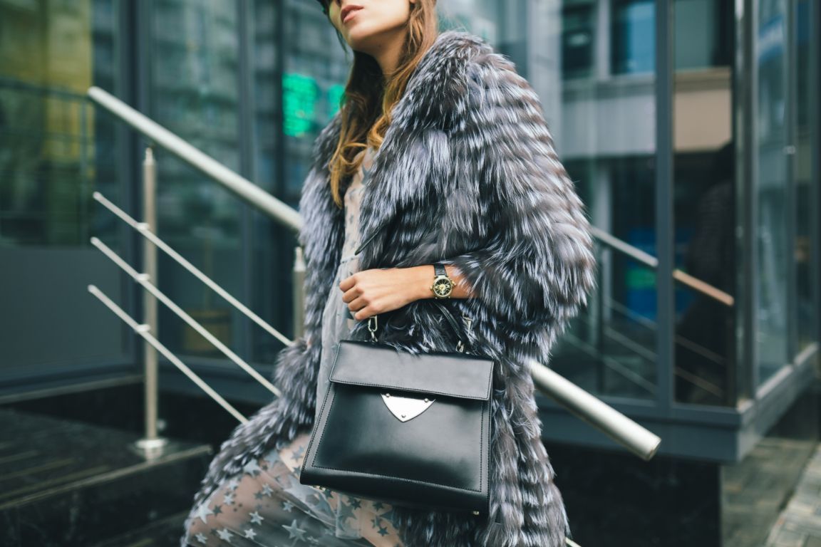 Una persona con un cappotto di pelliccia grigio e una borsa nera davanti a un edificio di vetro.