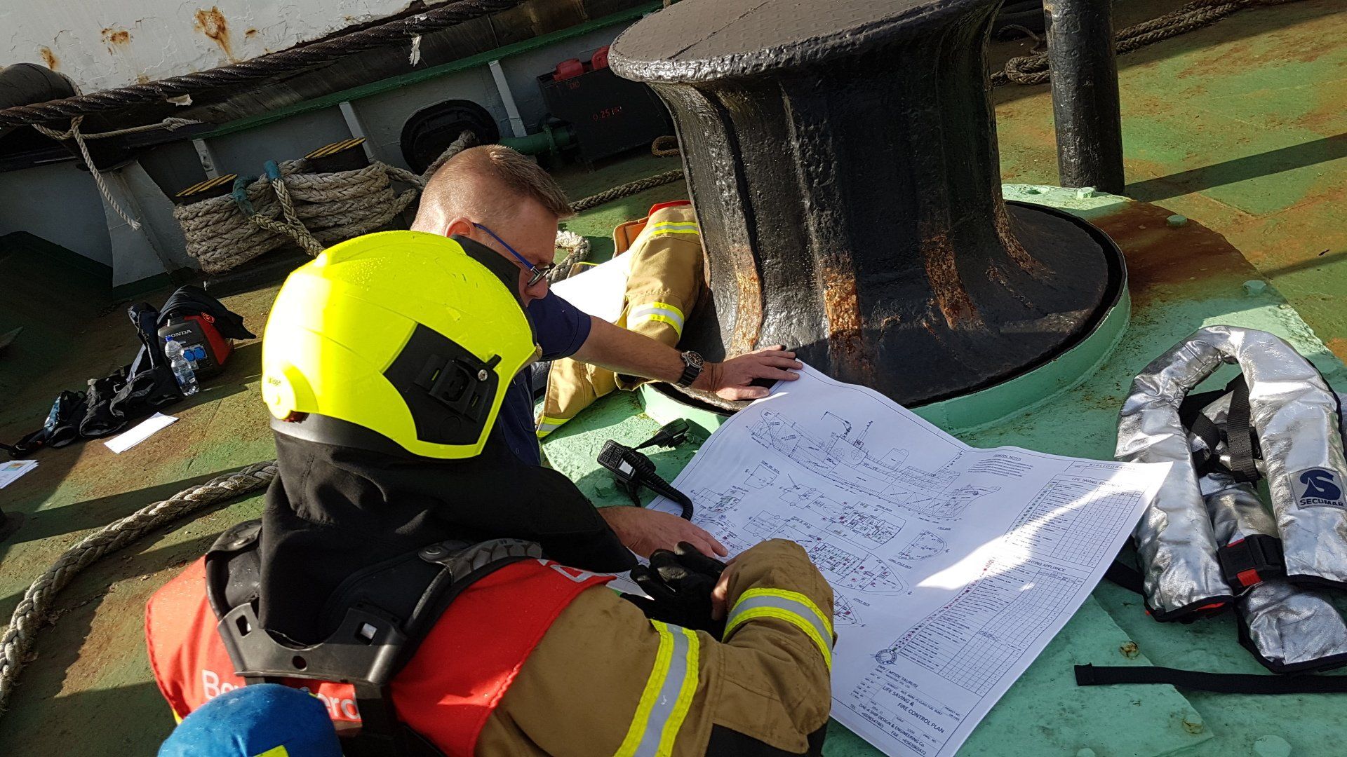 Twee mensen in brandweerkleding bekijken een groot document op het dek van een schip, vlakbij een groot metalen voorwerp.