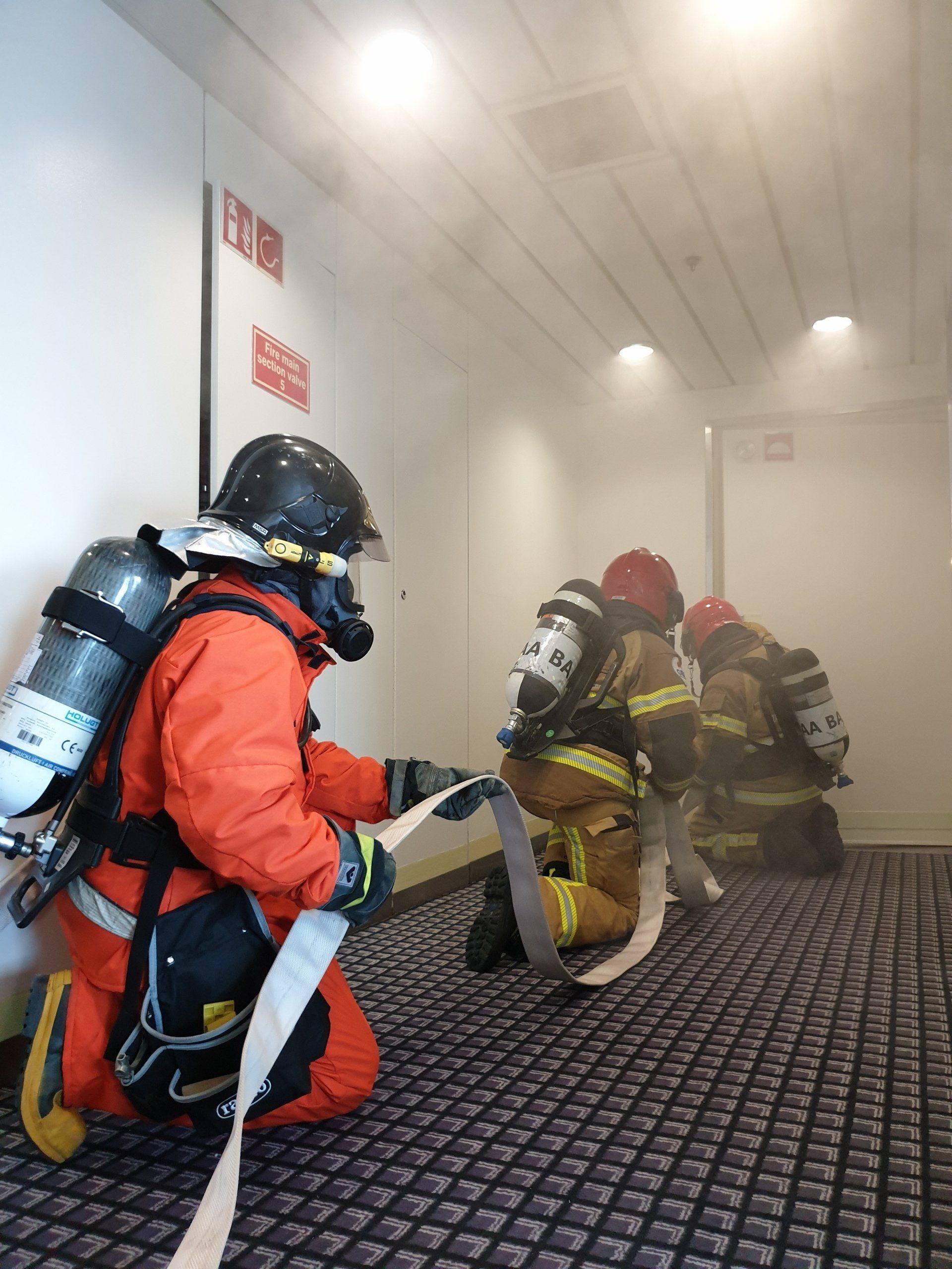 Brandweerlieden in beschermende kleding vechten in de met rook gevulde gang. Eén knielt met een slang in de hand. 