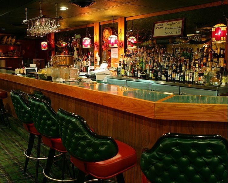 Dimly lit bar interior with a long wooden counter, green stools, and rows of bottles behind the bar