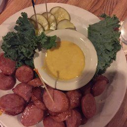 Plate of sliced sausage with kale, pickles, and a bowl of yellow mustard on a wooden table