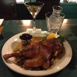 Roasted duck on a plate with citrus garnish, sauce, a martini, and water on a restaurant table