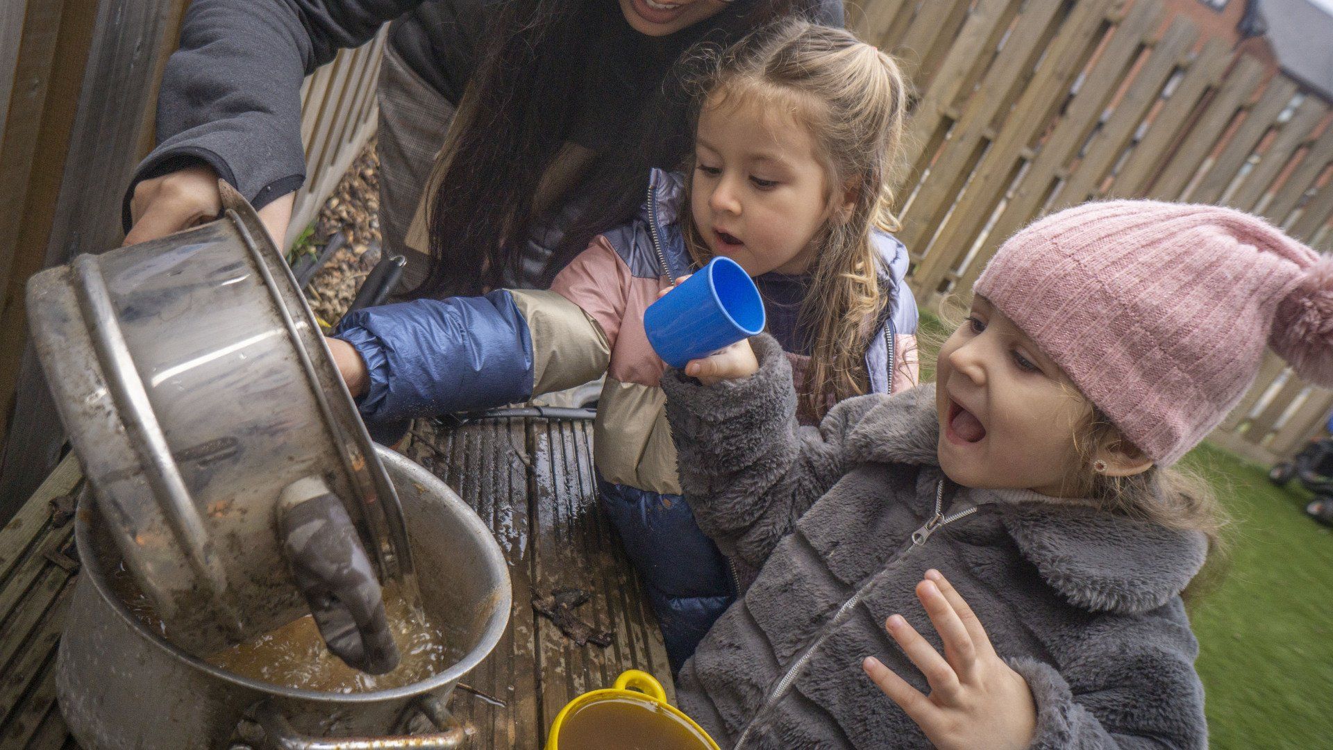 Two girls in jackets watch as someone pours liquid from a pot. One girl holds a blue cup, the other wears a pink hat.