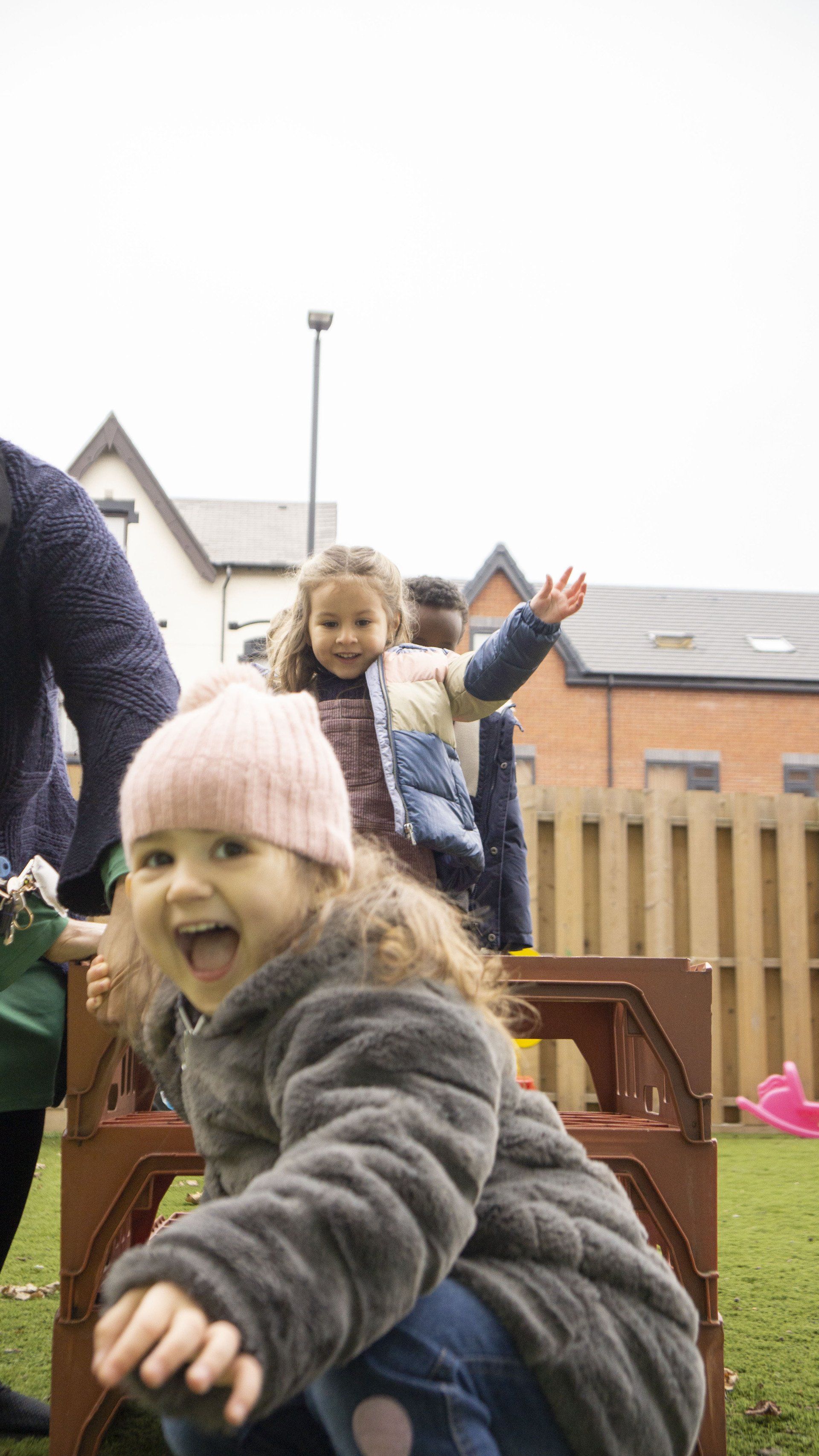 Children playing outside, one smiling and reaching, near a wooden fence.
