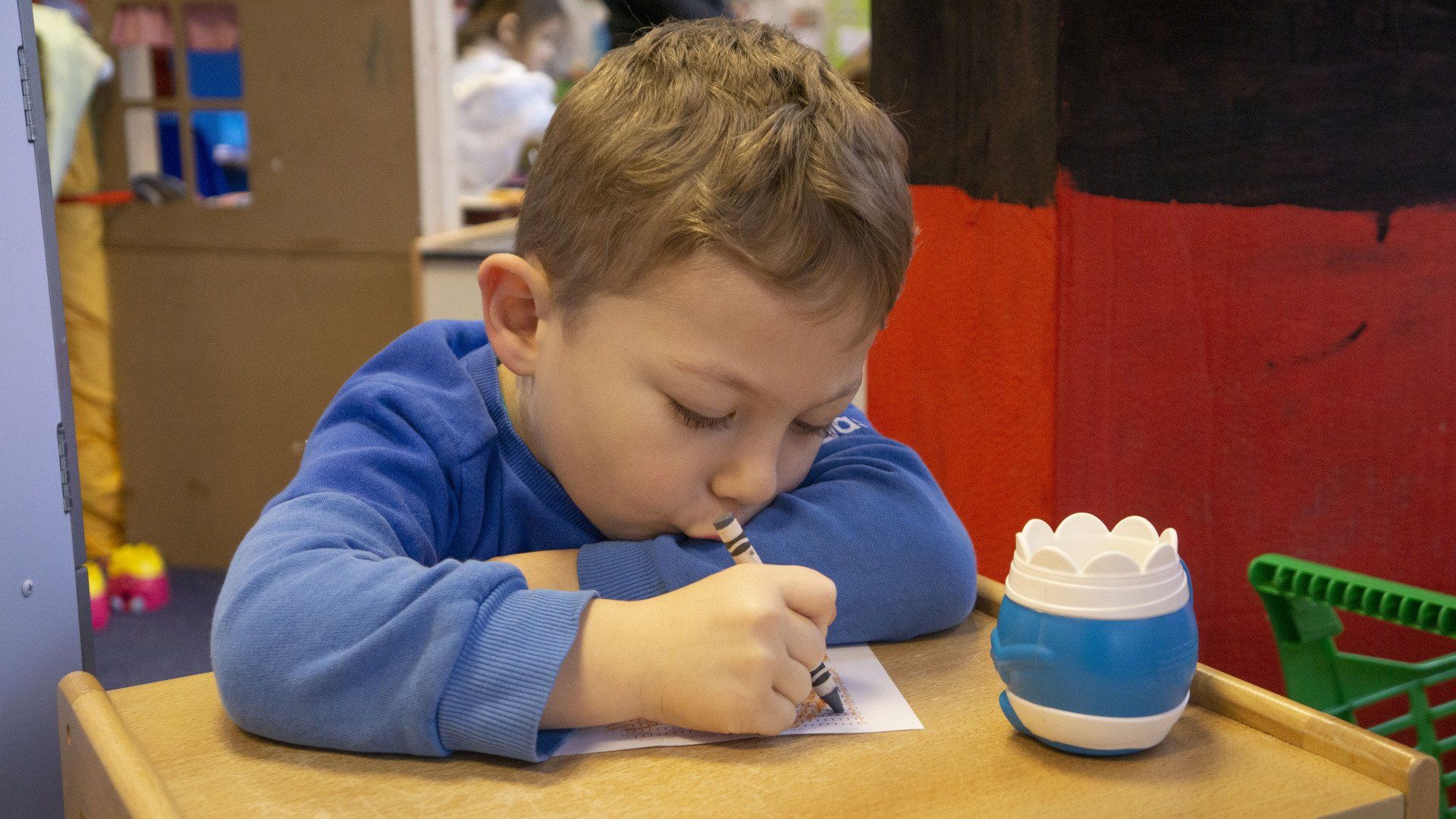 Boy in blue shirt drawing at a desk, with a blue and white container.