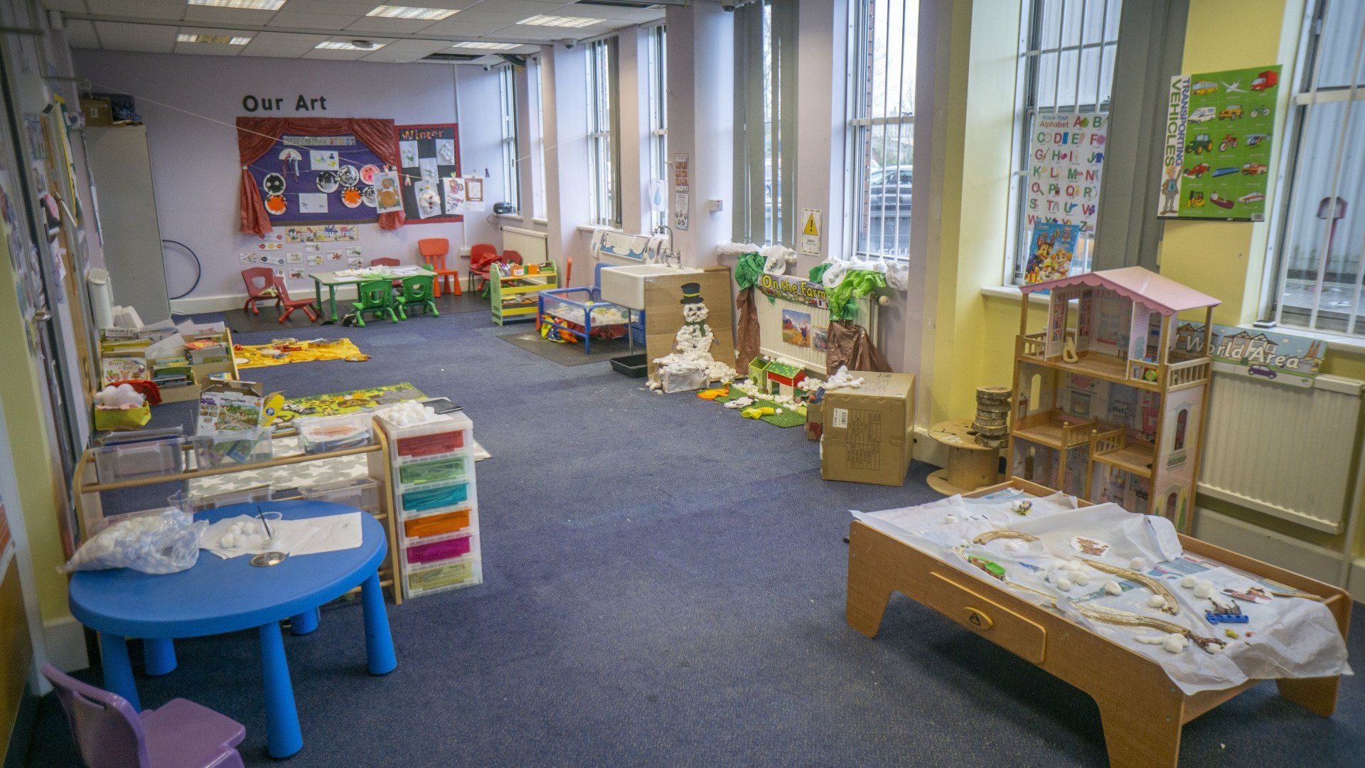 Childcare classroom, toys, tables, and artwork on the walls. Blue carpet, natural light.