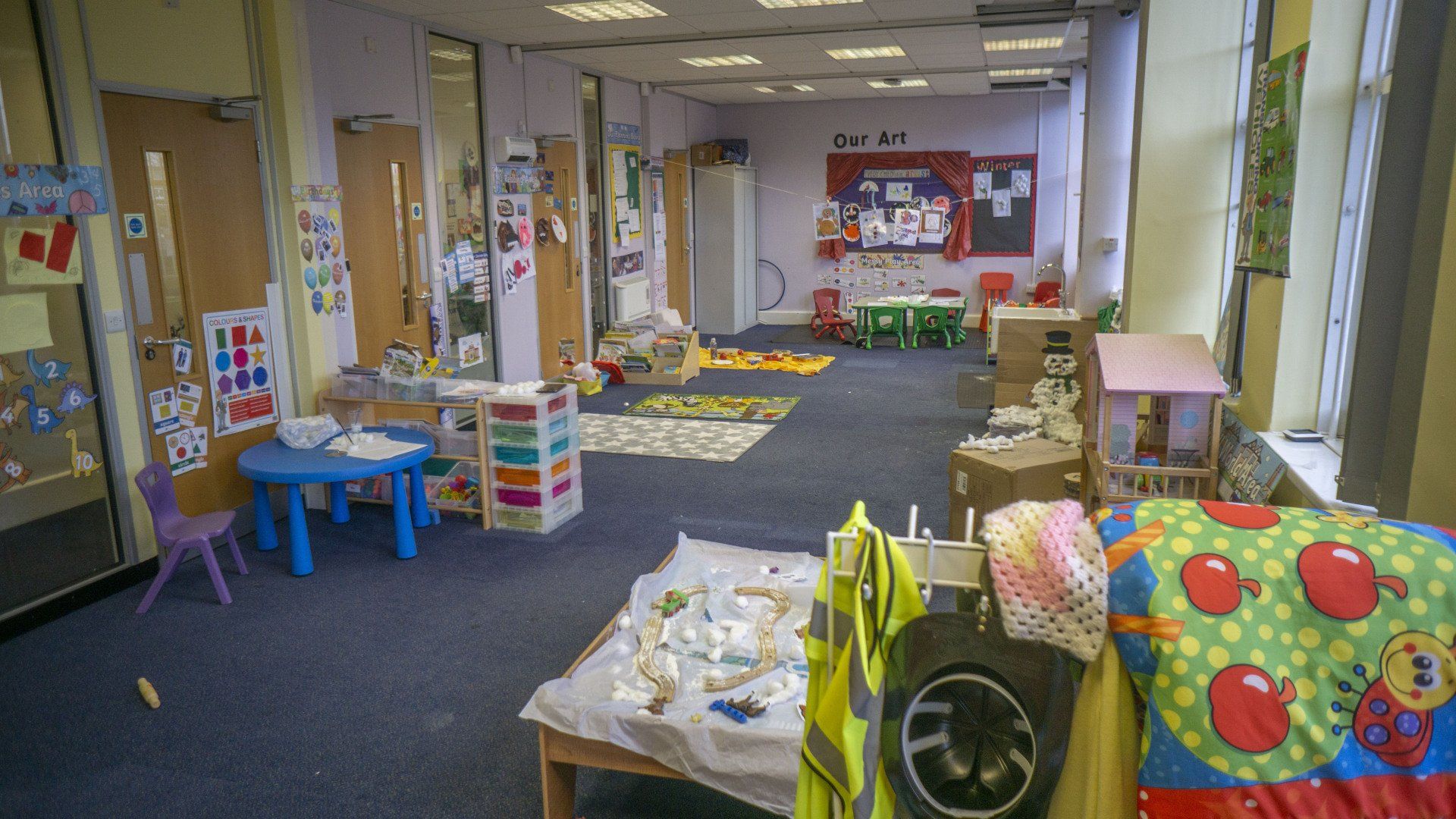 Playroom with tables, toys, and child-sized furniture. Blue carpet, light walls, and several doors visible.