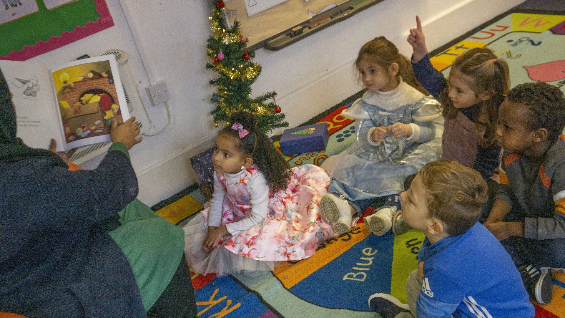 A teacher reads a book to a group of children in a classroom with a Christmas tree.