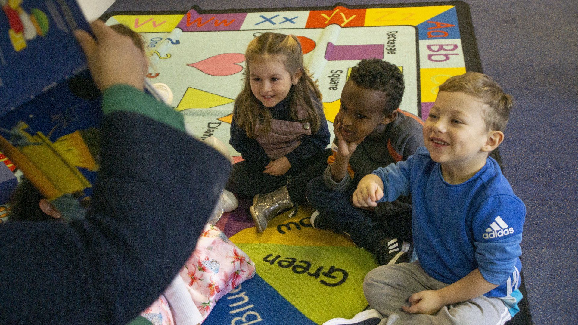 Children sitting on a rug, listening to a person reading a book. One child points, smiling.