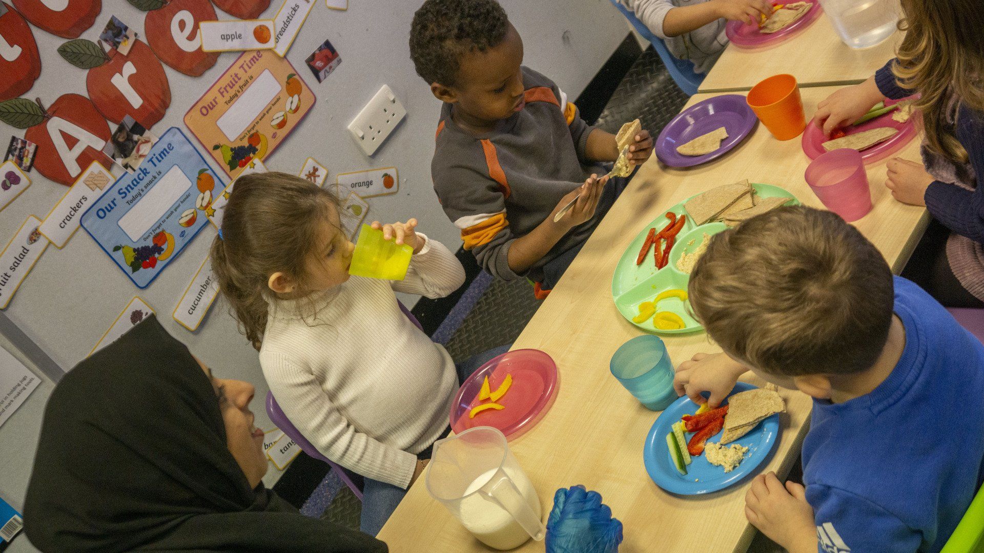 Children and adult eating at a table, snacks and drinks are present. Interior setting.
