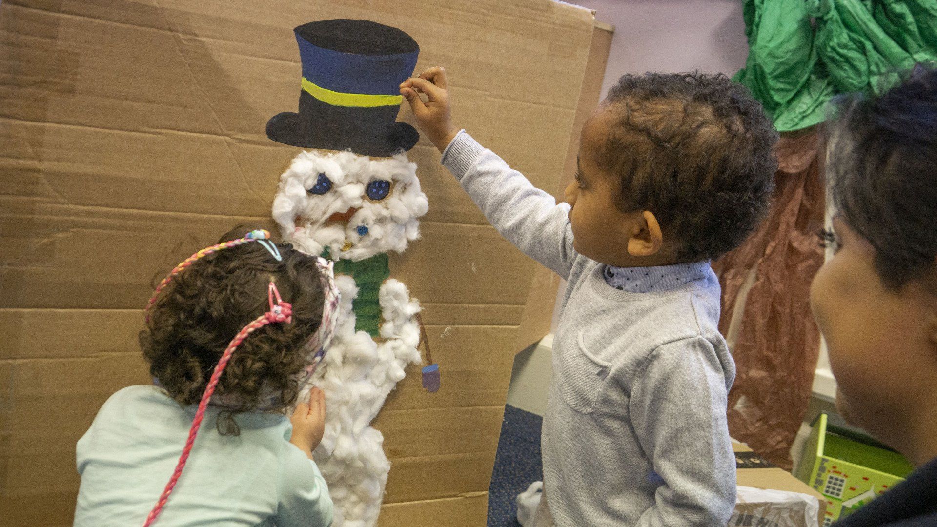 Children decorate a snowman made of cardboard and cotton against a wood-paneled wall.