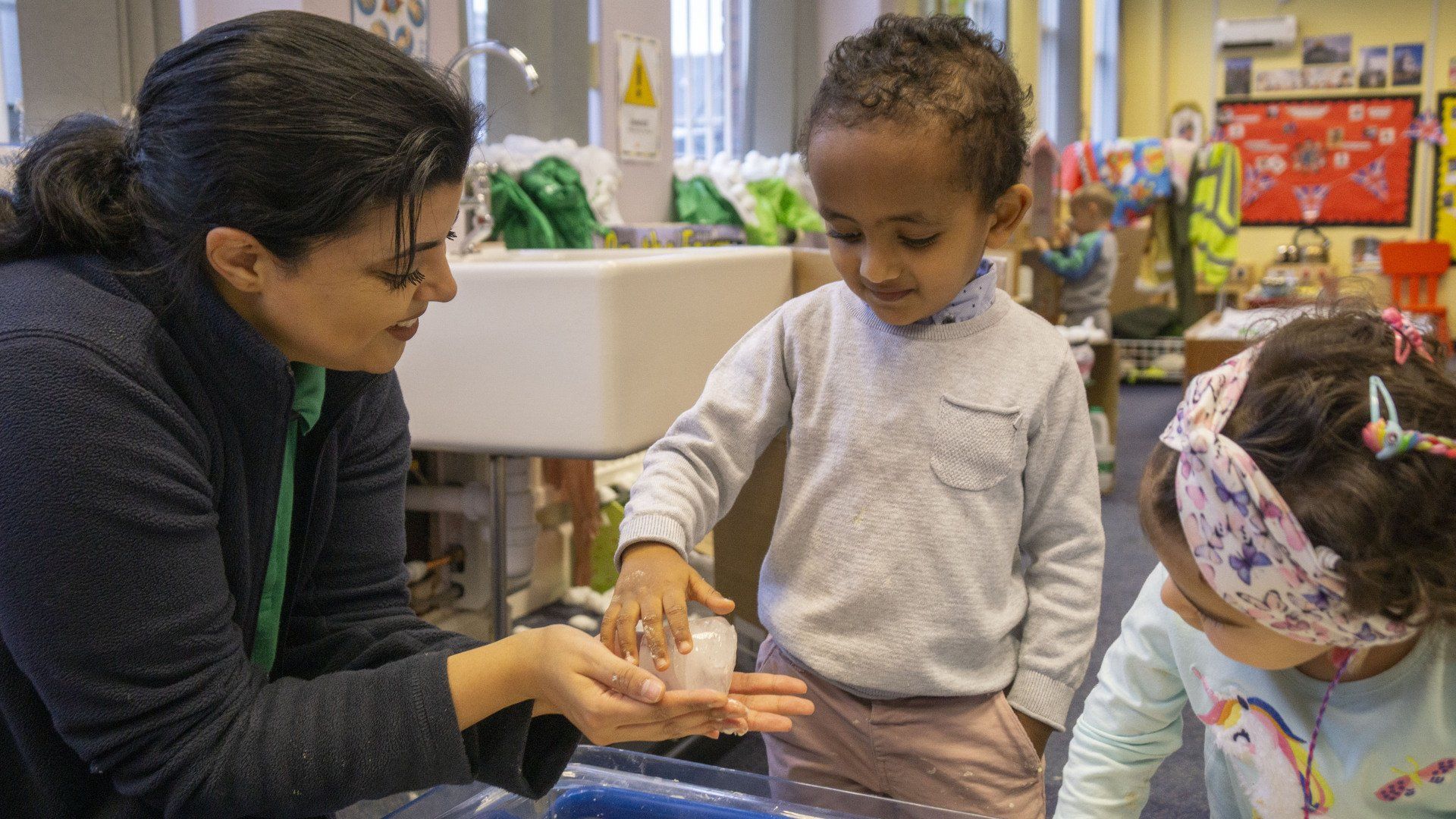 Teacher helping a child wash hands at a sink. A second child watches.