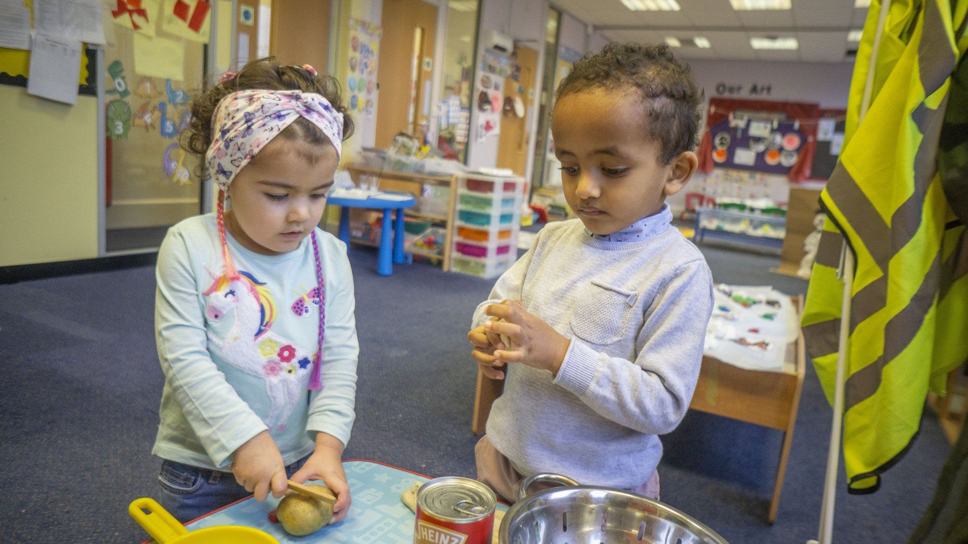 Two children peeling a potato at a table in a brightly lit preschool classroom.