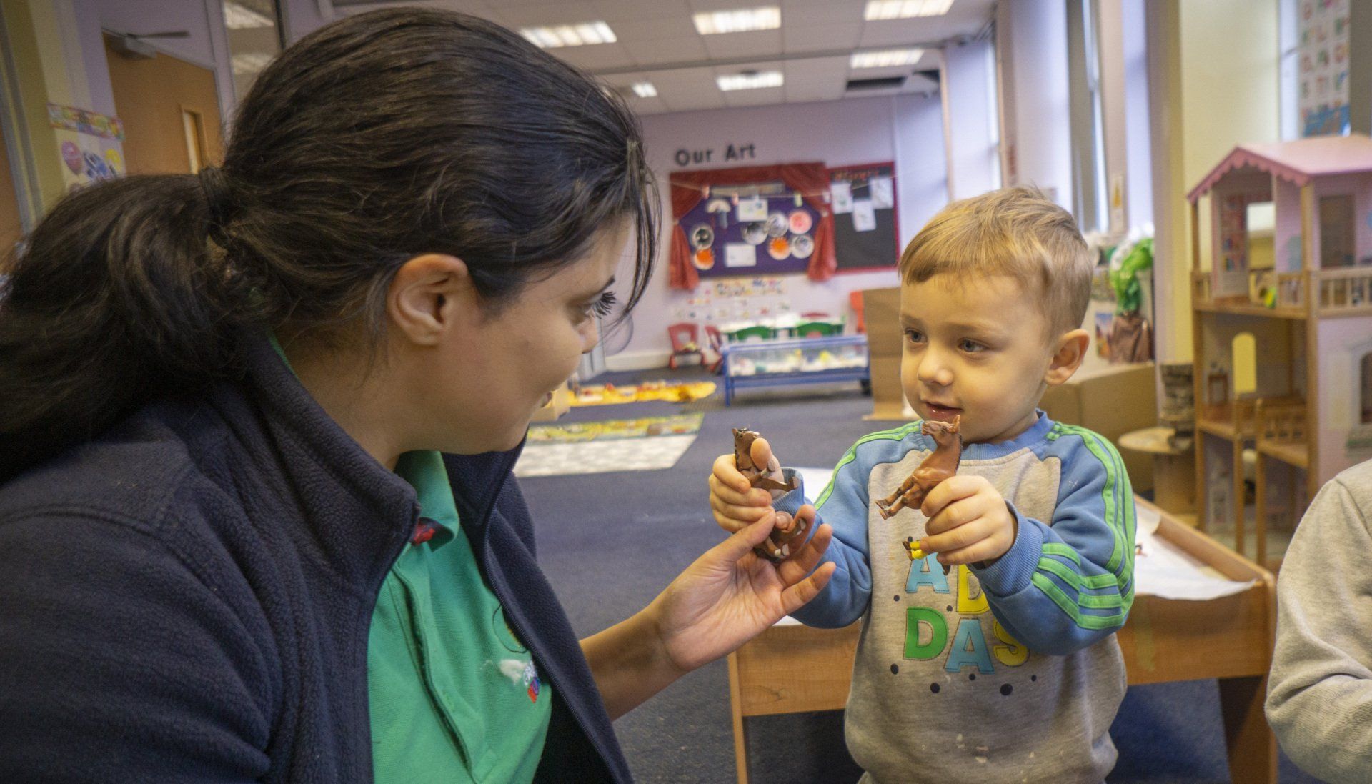 A person and a toddler playing with toy figurines in a classroom setting.