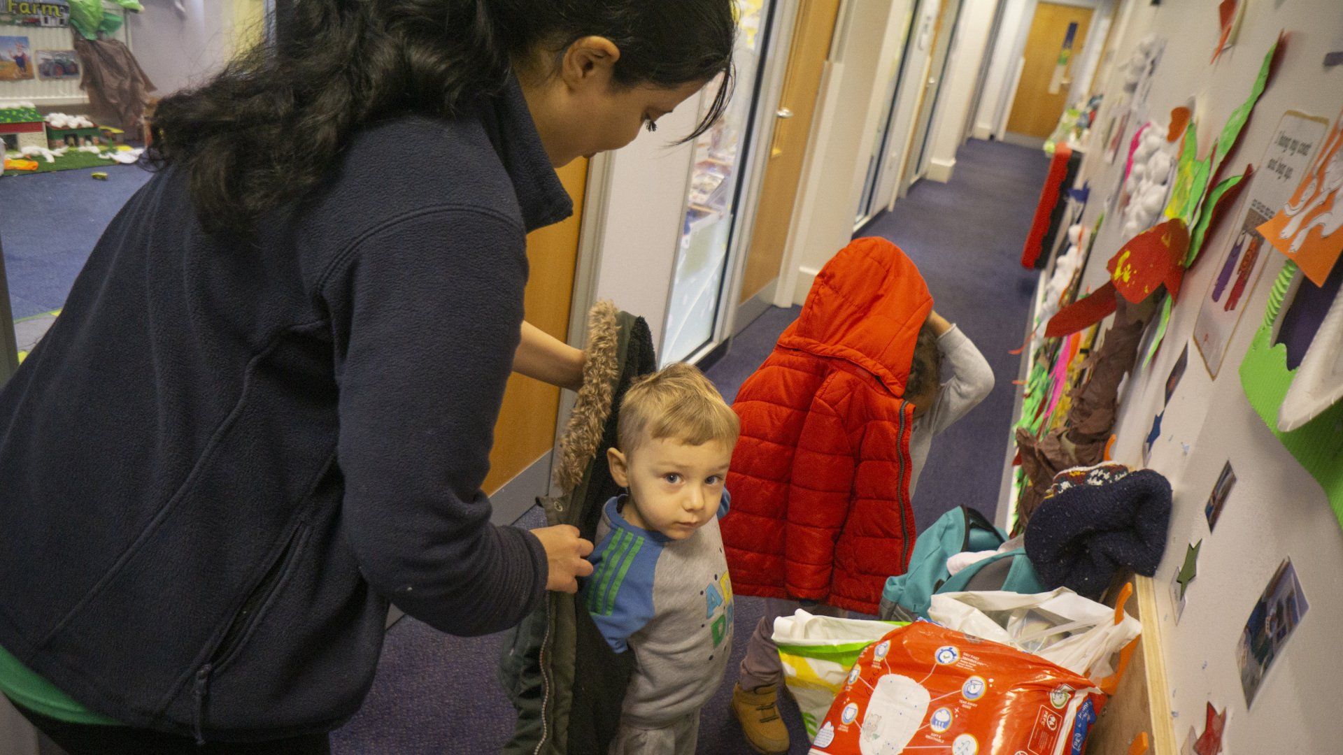 Woman helping a child with a coat in a hallway with coat rack.