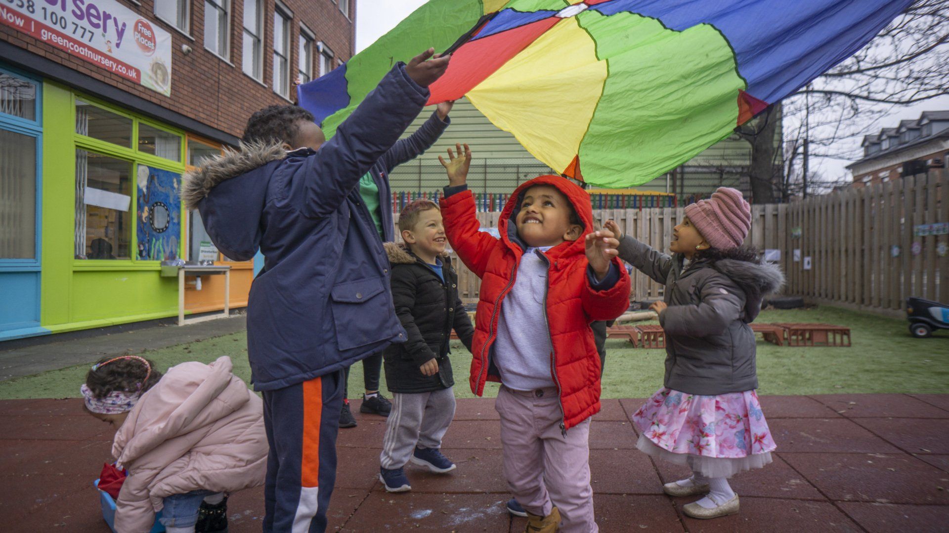 Children playing with a colorful parachute outside a nursery.