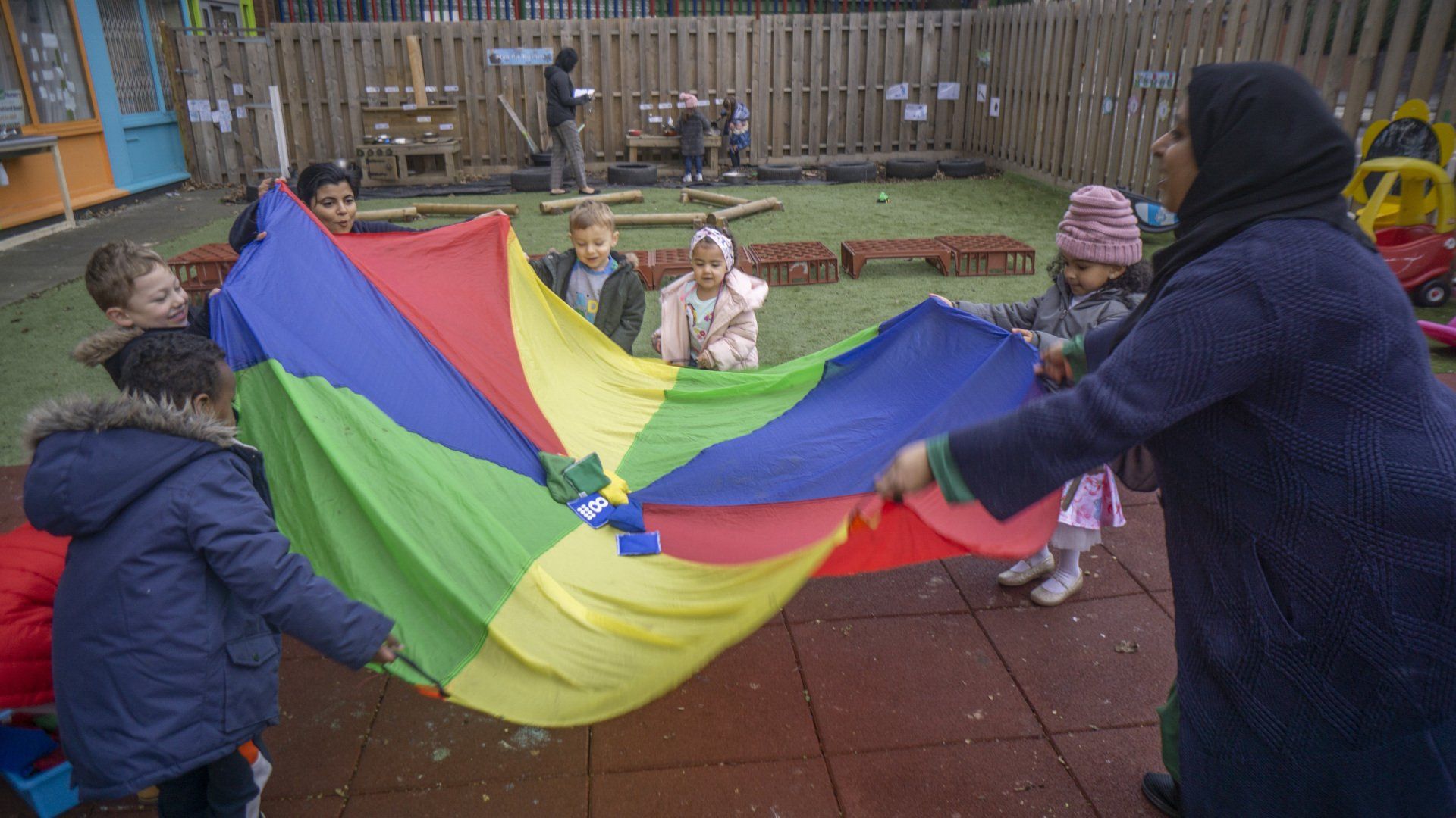 Children and an adult playing with a rainbow parachute in an outdoor setting.