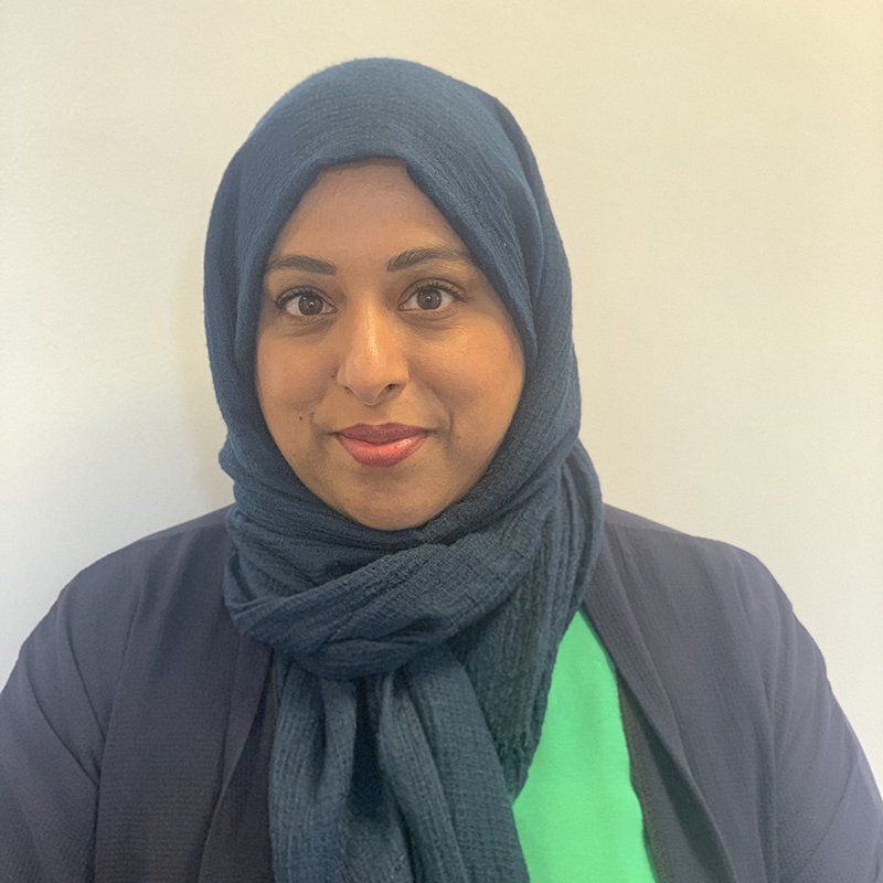 Woman wearing a blue hijab, navy blazer, and green top smiles at the camera against a white background.