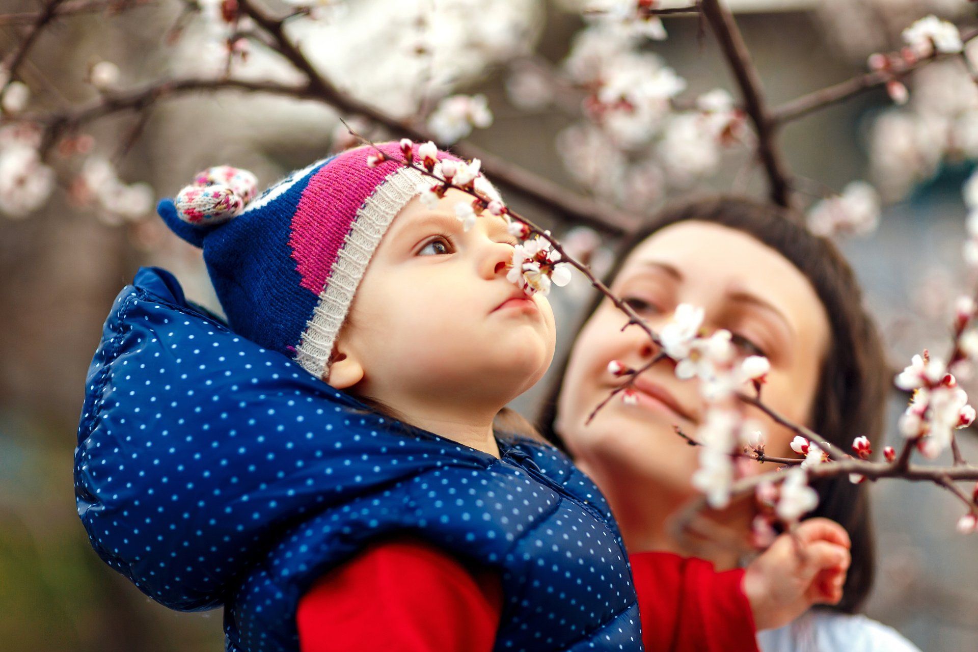 Child in blue jacket and hat smelling flowers, with a woman in background.