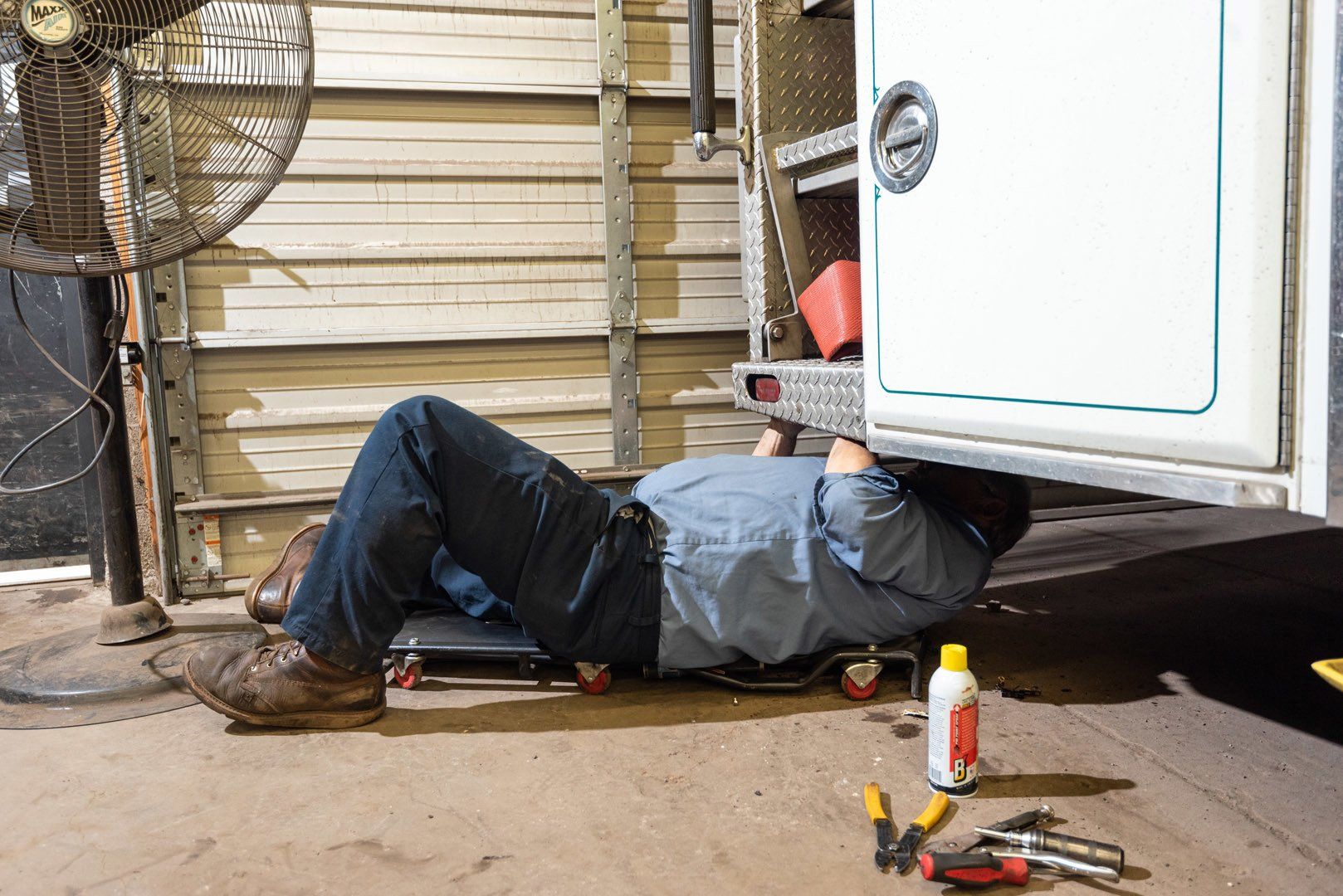 Mechanic on a creeper working under a white truck in a garage. Tools and spray can on the floor.