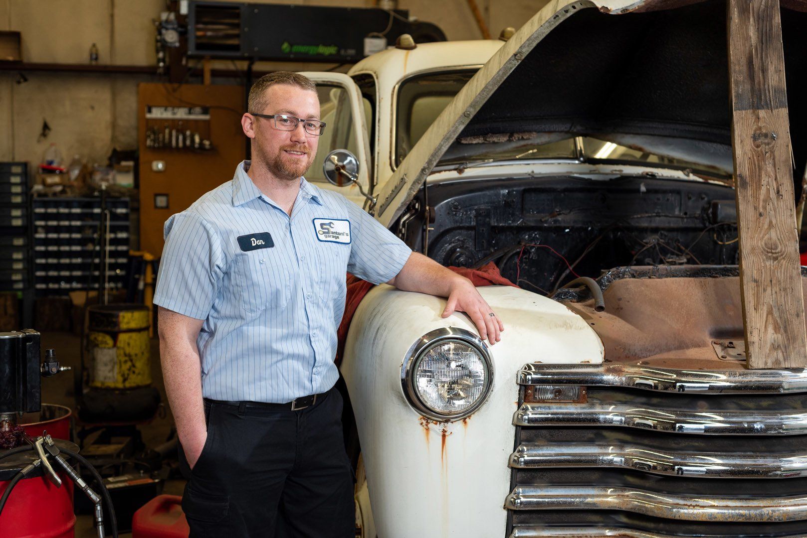 Mechanic in a blue shirt stands by a vintage white truck with the hood up in a garage.