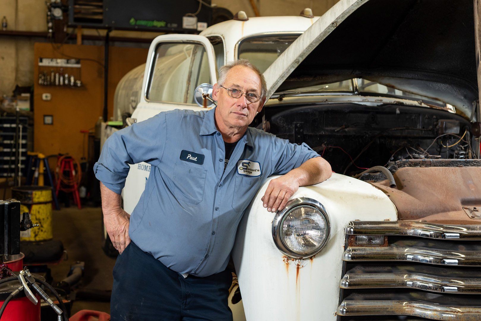 Mechanic in blue shirt leans on classic car in a garage with open hood.