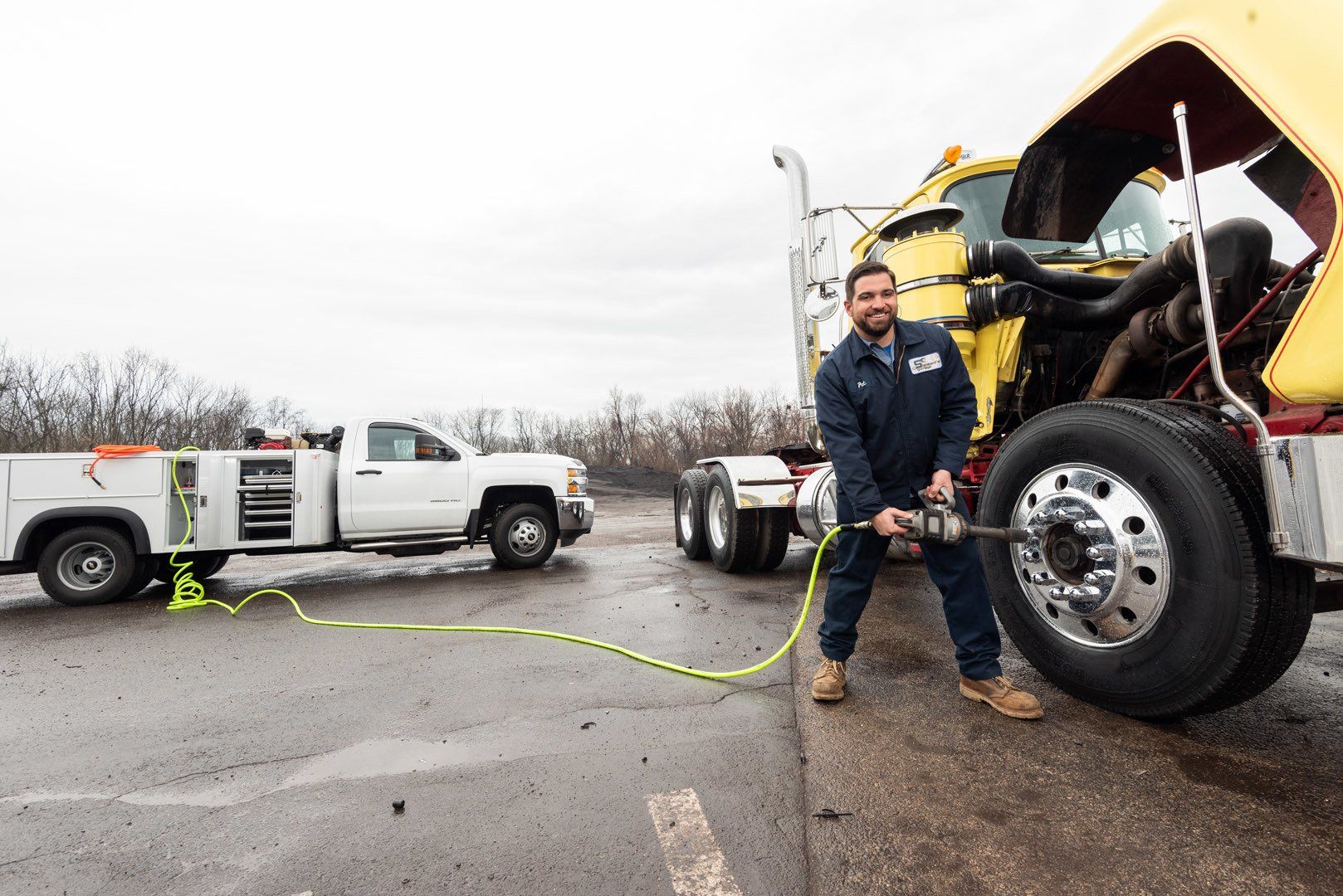 Mechanic using an impact wrench on a semi-truck tire, with a work truck in the background on a wet, overcast day.
