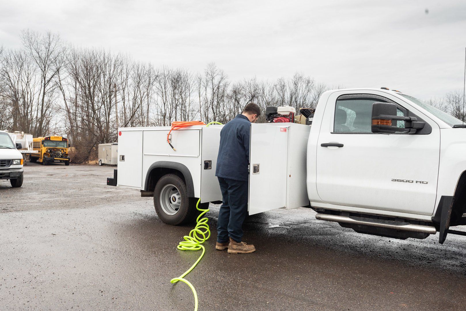 Man in a blue jacket opens a white truck toolbox on a pickup truck. A yellow hose lies on the asphalt.