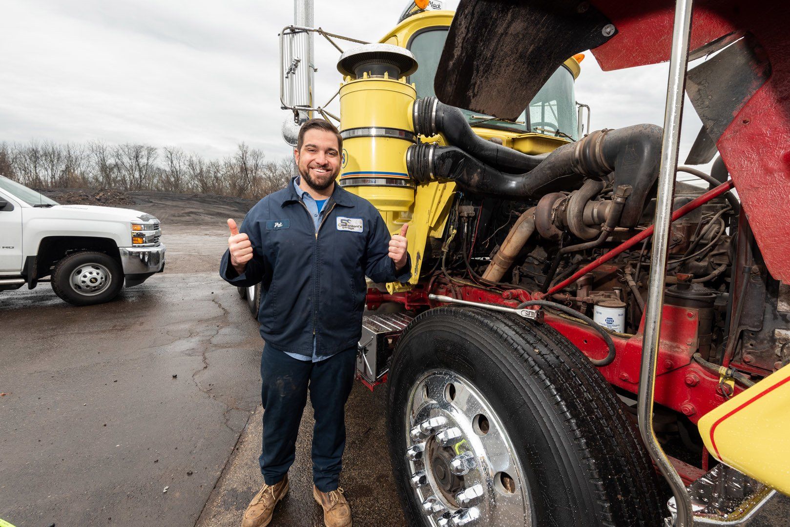 Mechanic with thumbs-up in front of a yellow semi-truck with open hood. Outdoors, gray day.