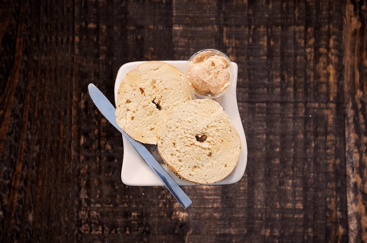 Two bagel halves and a small portion of spread on a white plate with a knife, set on a rustic dark wooden surface.