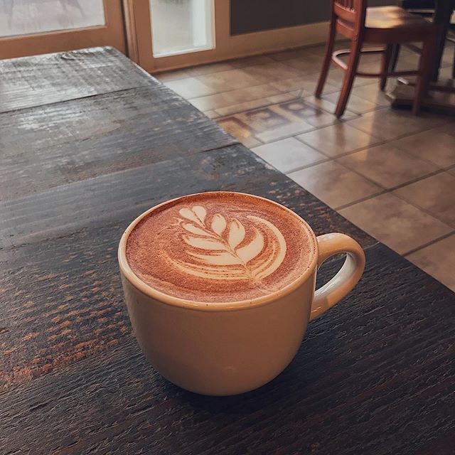 A ceramic mug of coffee with latte art in the shape of a leaf, sitting on a weathered wooden table in a cafe.