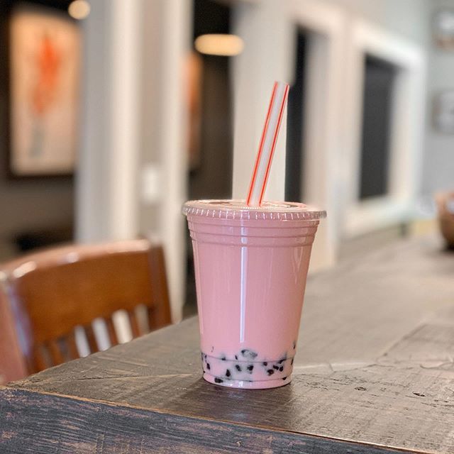 A pink bubble tea in a clear plastic cup with a striped straw, sitting on a wooden surface in a softly lit room.f