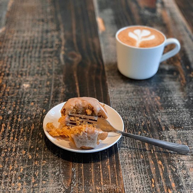 A partially eaten glazed muffin on a white saucer with a fork next to a mug of coffee on a dark wooden table.