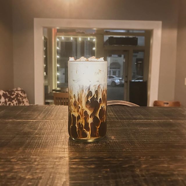 An iced latte with a thick, white foam layer settling into dark coffee, sitting on a wooden table in a dimly lit cafe.