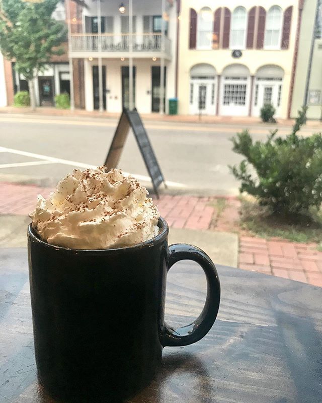A black mug topped with whipped cream and cocoa powder sits on a wooden table, overlooking a street with historic buildings.